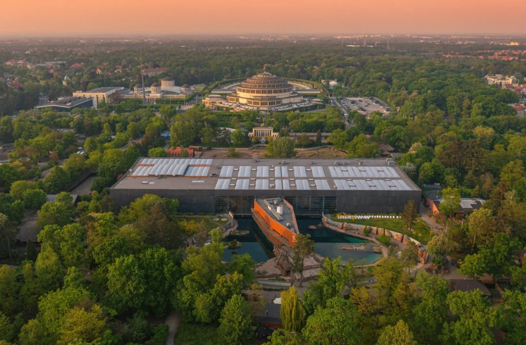 Widok z lotu ptaka na nowoczesny budynek Afrykarium w Zoo Wrocław oraz historyczną Halę Stulecia w tle o zachodzie słońca.