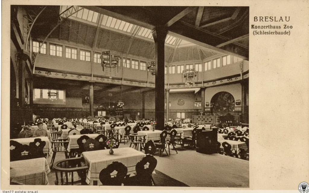 Stara pocztówka przedstawiająca wnętrze Konzerthaus Zoo (Schlesierbaude) we Wrocławiu (dawnym Breslau) z licznymi stolikami restauracyjnymi i ozdobnymi żyrandolami pod wysokim sufitem.