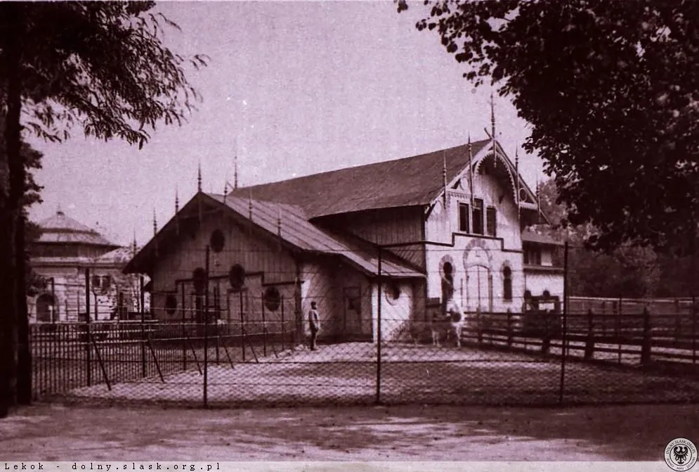 Zabytkowy drewniany budynek dawnej żyrafiarni w Ogrodzie Zoologicznym we Wrocławiu, przedstawiony na tle zieleni i ogrodzenia.