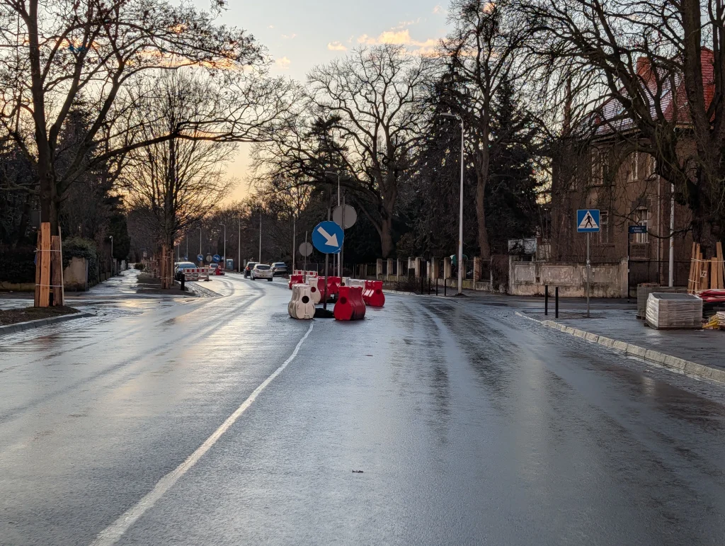 Mokra asfaltowa jezdnia o zmierzchu z ustawionymi barierami drogowymi oraz niebieskim znakiem nakazu jazdy w prawo.