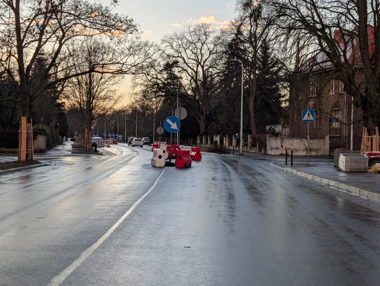 Mokra asfaltowa jezdnia o zmierzchu z ustawionymi barierami drogowymi oraz niebieskim znakiem nakazu jazdy w prawo.