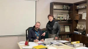 Uśmiechnięty mężczyzna z medalem na szyi podpisuje dokumenty przy biurku w obecności kobiety wskazującej na laptopa.