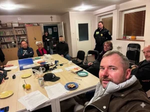 Grupa ludzi zgromadzona wokół długiego stołu w pokoju. Niektórzy siedzą, inni stoją, a na stole leżą dokumenty i drobne przedmioty. W tle widoczne regały z książkami i drzwi. Osoba na pierwszym planie patrzy w kierunku kamery.