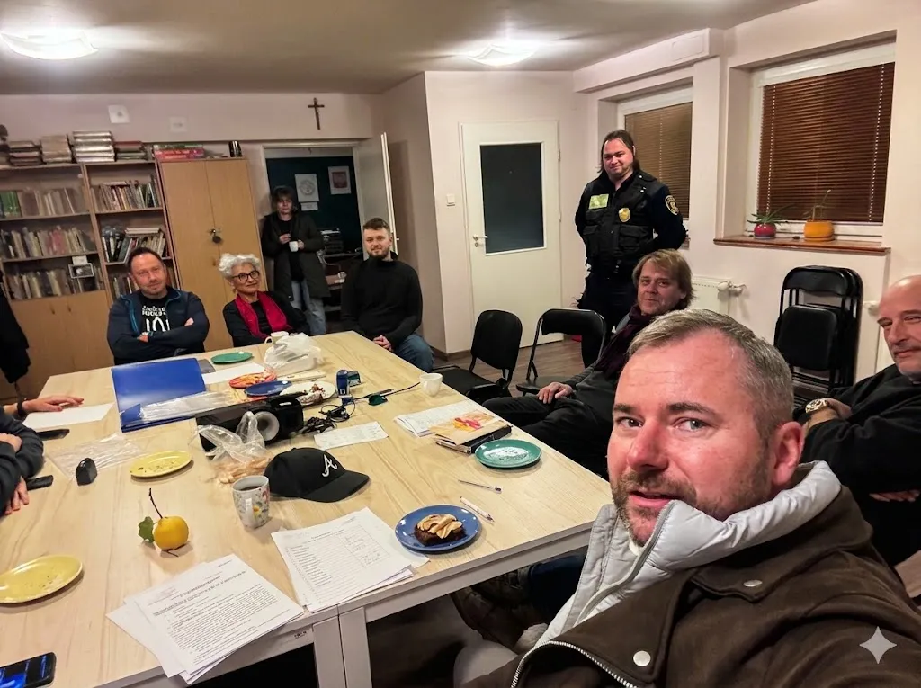 Grupa ludzi zgromadzona wokół długiego stołu w pokoju. Niektórzy siedzą, inni stoją, a na stole leżą dokumenty i drobne przedmioty. W tle widoczne regały z książkami i drzwi. Osoba na pierwszym planie patrzy w kierunku kamery.